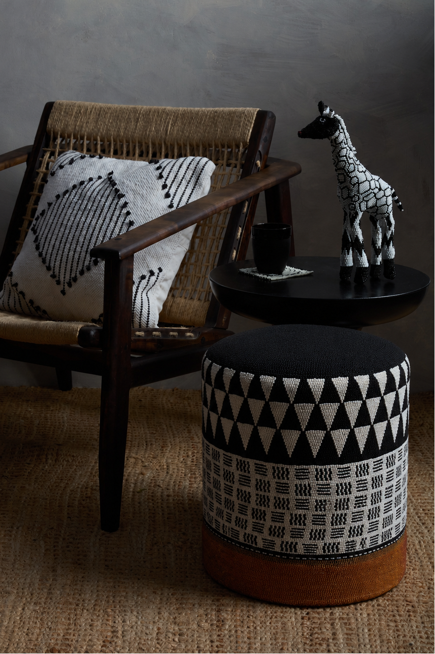 A black and white beaded patterned pouf ottoman with a brown leather base sits next to a mid-century modern armchair with a woven seat and a matching throw pillow. A small black side table holds a black cup and a decorative giraffe figurine against a dark gray wall.