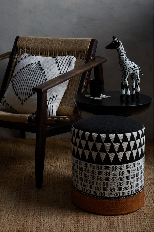A black and white beaded patterned pouf ottoman with a brown leather base sits next to a mid-century modern armchair with a woven seat and a matching throw pillow. A small black side table holds a black cup and a decorative giraffe figurine against a dark gray wall.