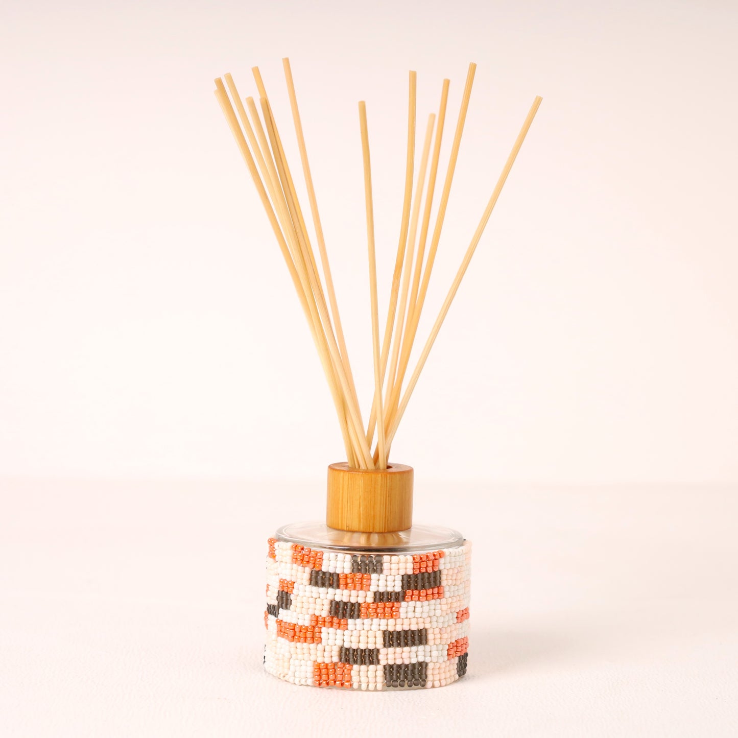 A reed diffuser with a cylindrical glass bottle wrapped in a white, orange, and black geometric bead pattern, topped with a light wood collar and natural bamboo reeds fanned out against a clean white background.