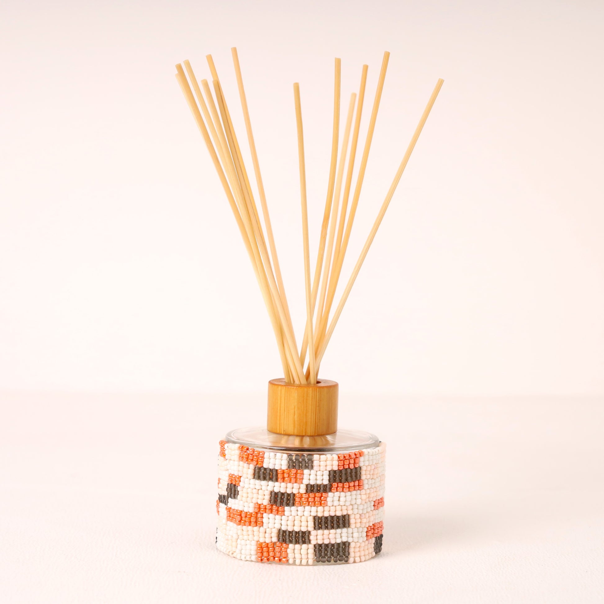 A reed diffuser with a cylindrical glass bottle wrapped in a white, orange, and black geometric bead pattern, topped with a light wood collar and natural bamboo reeds fanned out against a clean white background.