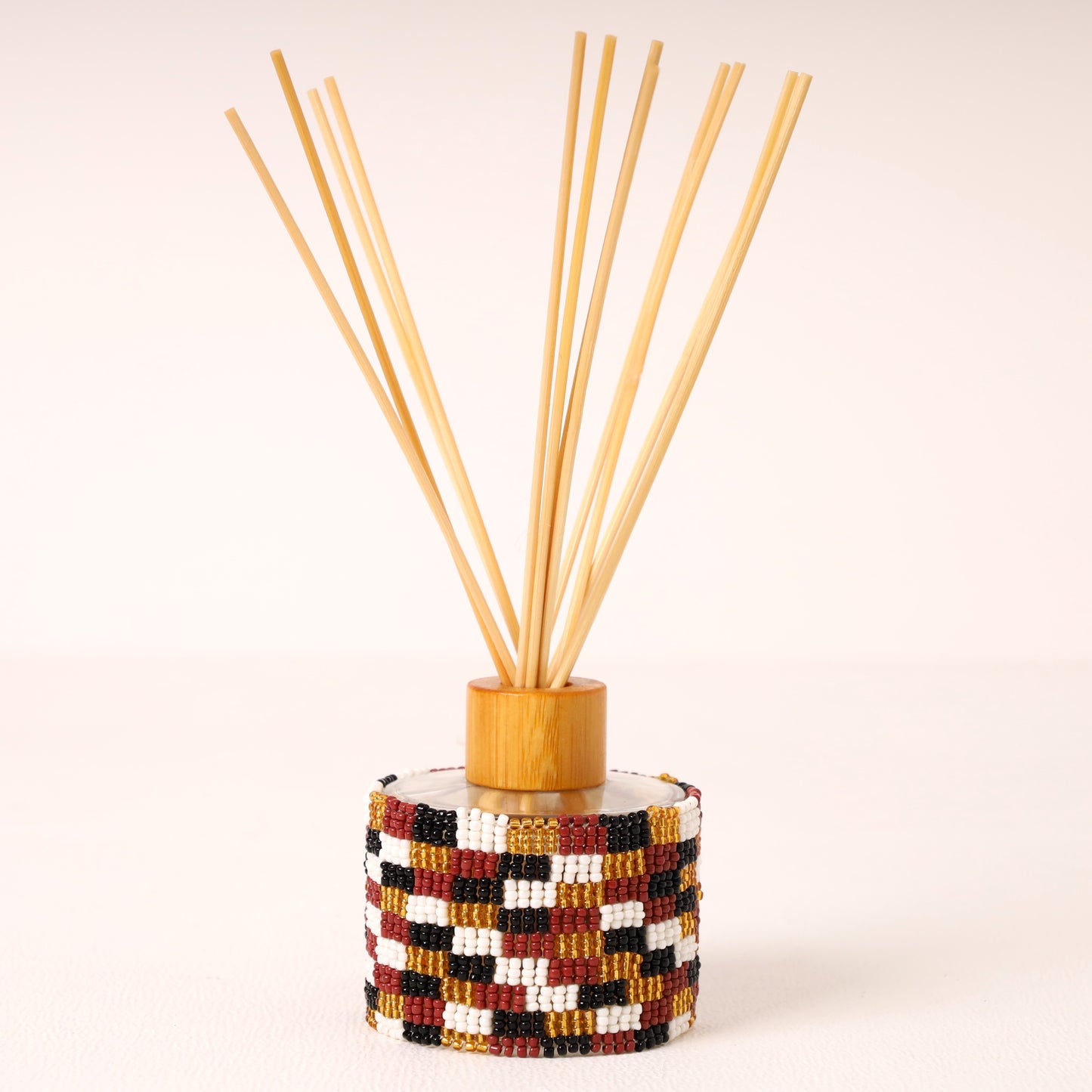 A reed diffuser with a cylindrical base wrapped in a geometric bead pattern of red, black, white, and gold, topped with a light wood collar and natural bamboo reeds fanned out against a warm off-white background.