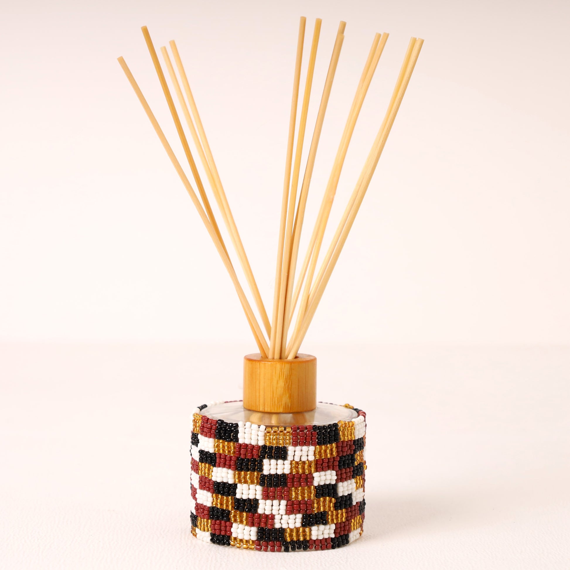A reed diffuser with a cylindrical base wrapped in a geometric bead pattern of red, black, white, and gold, topped with a light wood collar and natural bamboo reeds fanned out against a warm off-white background.