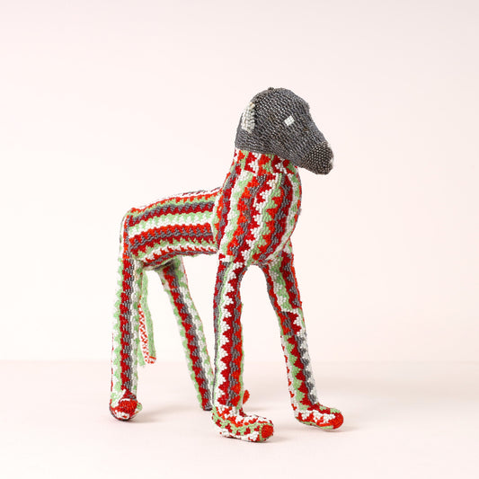 A handmade beaded baboon sculpture, featuring a gray head and legs with a body covered in red, green, and white textured stripes, set against a plain light background.