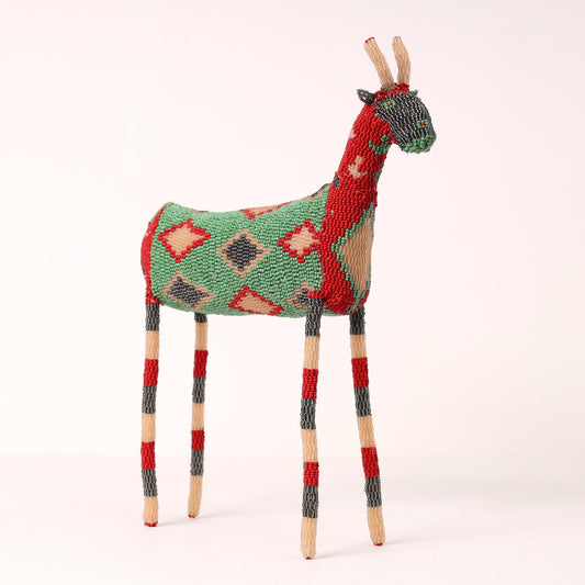 A handmade beaded springbok figurine with a green, red, and beige diamond-patterned body, standing on four striped legs against a plain light background.