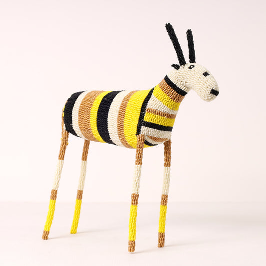 A handcrafted beaded antelope figurine with a striped body in yellow, black, brown, and cream, standing on a light beige background.