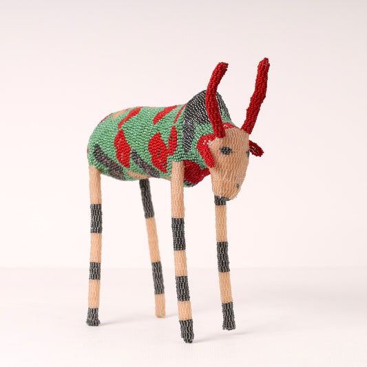 A handcrafted beaded figurine of a bull with red antlers, a green and red patterned body, and striped beige and black legs, set against a plain light background.