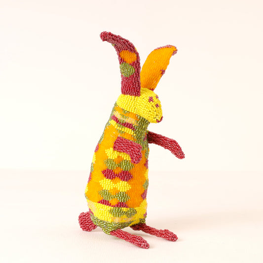 A colorful, handcrafted beaded figurine of a rabbit standing upright, featuring a yellow body with red, green, and orange patterns, set against a plain off-white background.