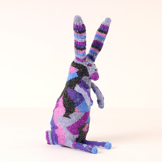A handmade beaded rabbit figurine in a sitting position, decorated with a colorful abstract pattern of purple, pink, blue, black, and gray beads on a plain light background.