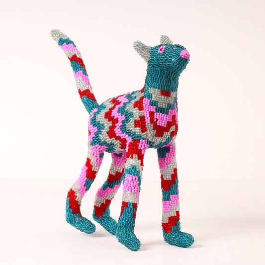 A handmade, beaded cat figurine in a standing pose, decorated with a colorful geometric pattern of teal, pink, red, and gray on a plain light background.