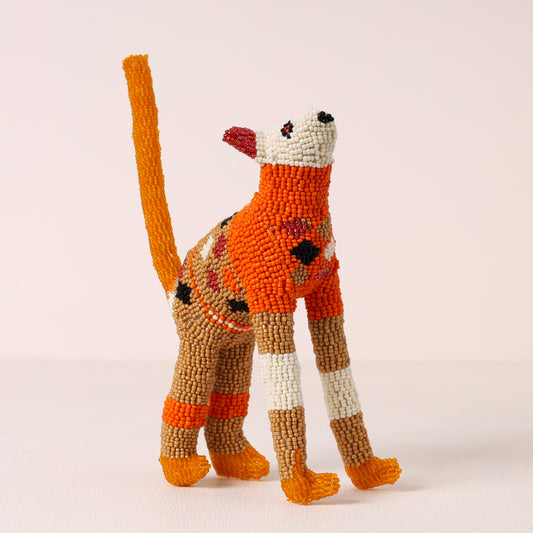 A handmade beaded cat figurine standing on a light beige background. The cat is primarily orange and brown with white, black, and red accents on its face, legs, and body.