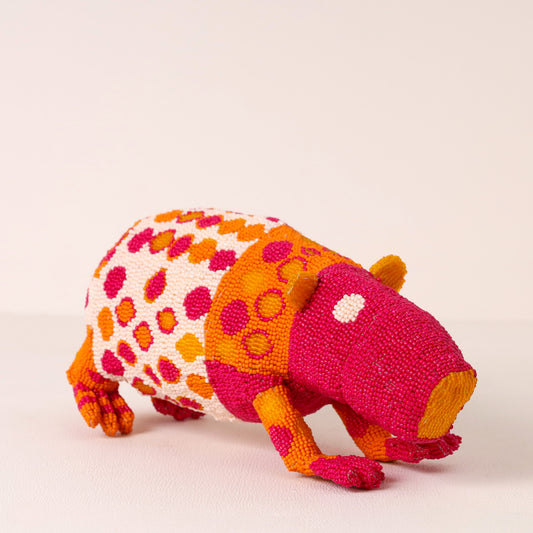 A handmade beaded dassie figurine, featuring a vibrant pattern of pink, orange, and yellow beads on a white base, set against a plain light beige background.