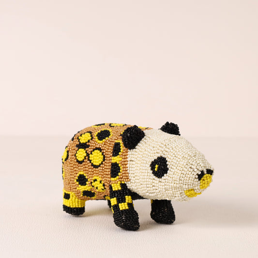 A small beaded dassie figurine with a white face, black ears and eyes, a yellow and black spotted body, and black legs, photographed against a plain light beige background.