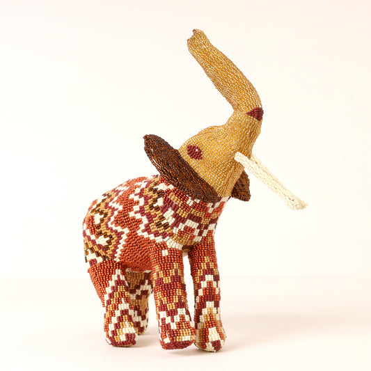 A handcrafted beaded elephant figurine with a tan trunk and tusks, featuring a geometric patterned body in red, brown, cream, and white, set against a plain off-white background.