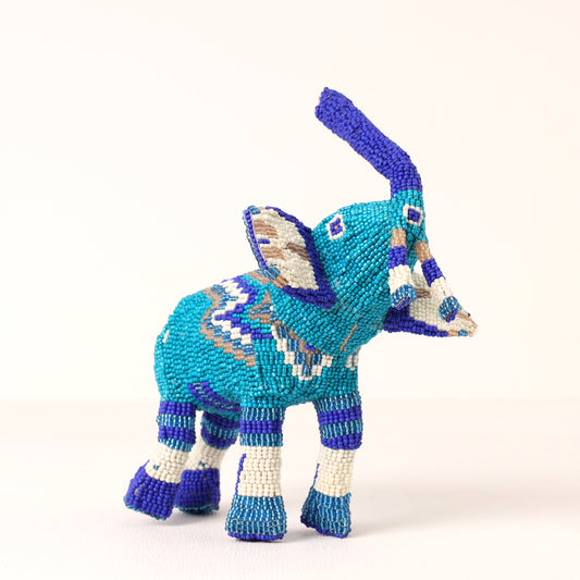 A handmade beaded elephant figurine in turquoise, blue, white, and purple, standing on a light cream background.