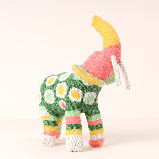 A colorful handmade beaded elephant sculpture with a green body, white and yellow spots, pink and green stripes on its legs, and a yellow and pink horn, photographed against a plain light background.