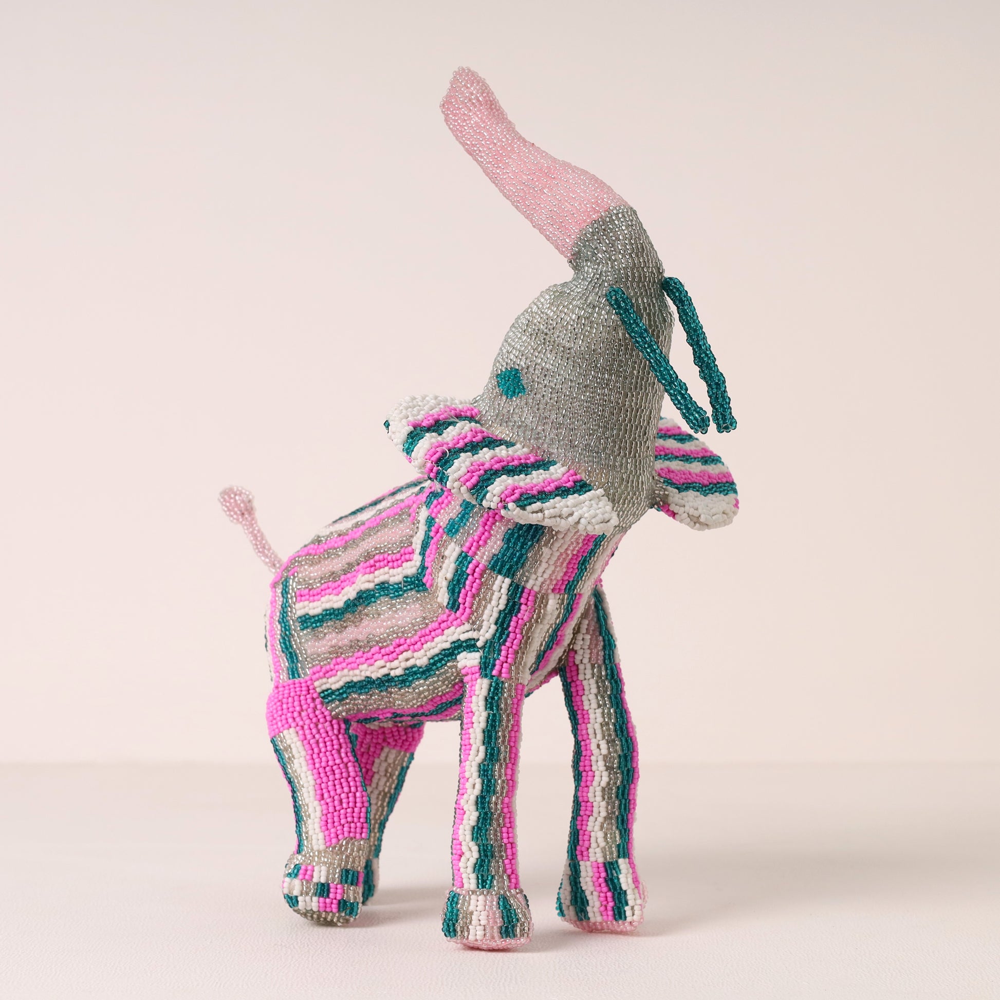 A handmade beaded elephant figurine, featuring a gray body, pink ears and tail, and pink, teal, and white striped body and legs, set against a solid pale pink background.
