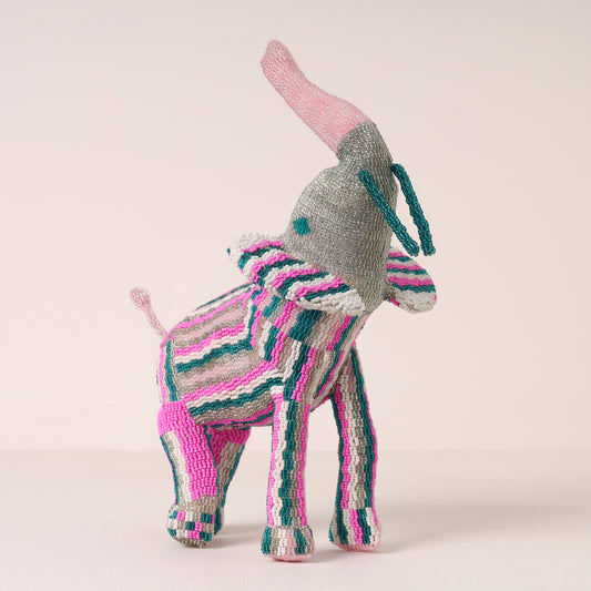A handmade beaded elephant figurine, featuring a gray body, pink ears and tail, and pink, teal, and white striped body and legs, set against a solid pale pink background.