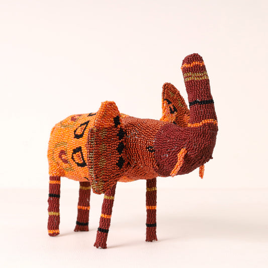 A handcrafted beaded elephant figurine, featuring a warm orange and reddish-brown body with black and yellow geometric patterns, set against a plain off-white background.