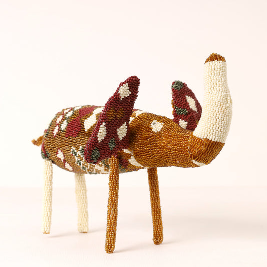 A handcrafted beaded elephant figurine featuring a brown body, cream-colored legs and tail, and reddish-brown ears with white and green bead accents, set against a plain light background.