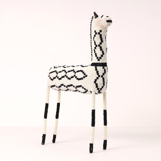 A handcrafted giraffe figurine made of white and black beads, standing on a light beige studio background.