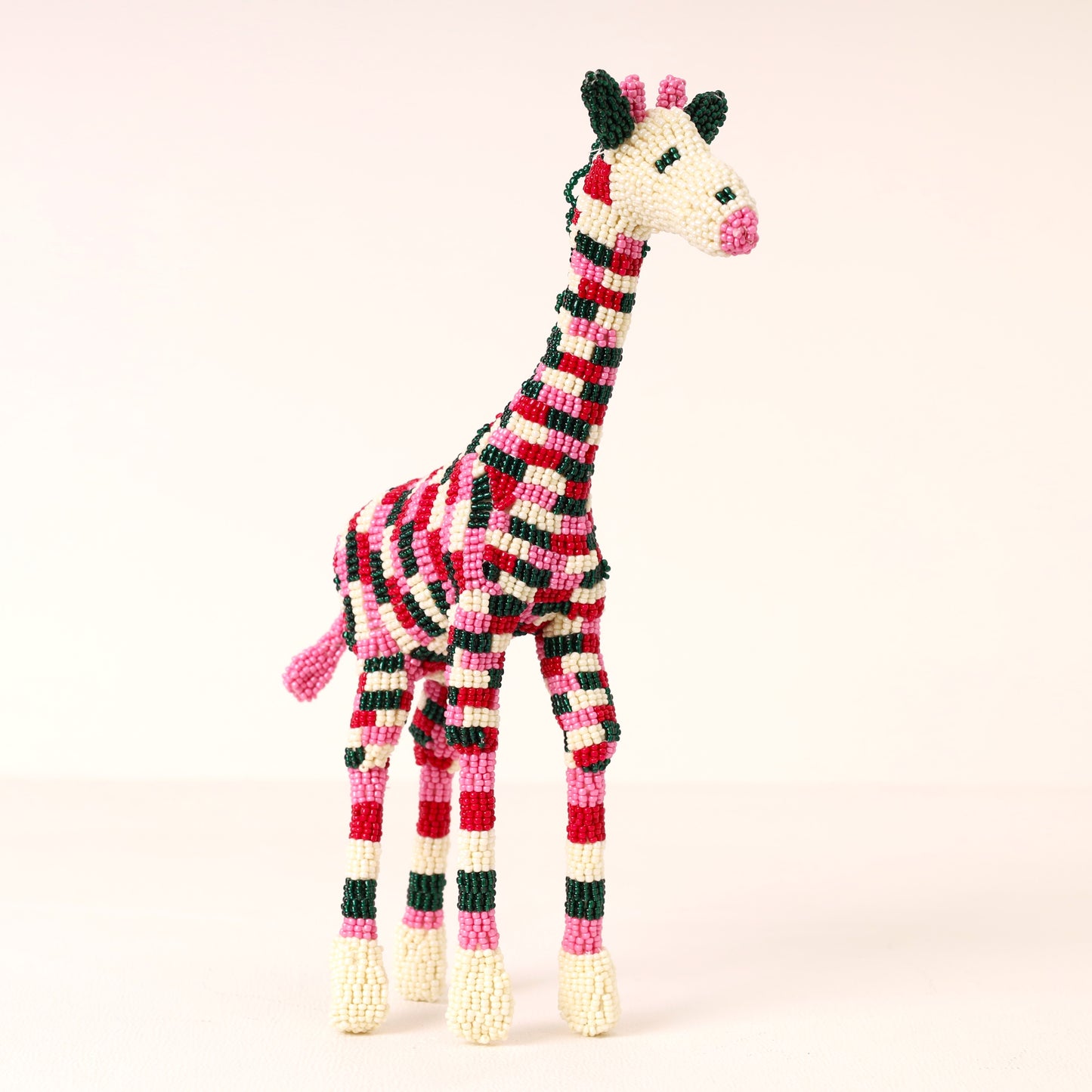 A handmade beaded giraffe sculpture with a red, black, and white striped pattern, pink accents, and cream-colored hooves, set against a solid pale pink background.