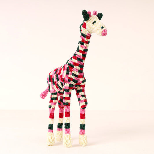 A handmade beaded giraffe sculpture with a red, black, and white striped pattern, pink accents, and cream-colored hooves, set against a solid pale pink background.