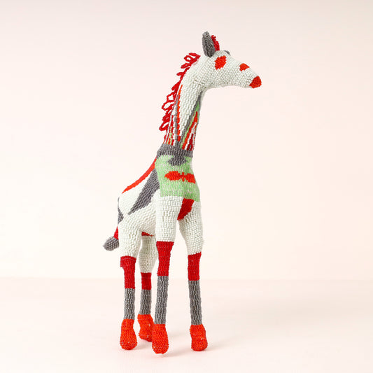 A hand-beaded giraffe sculpture with a white body, red and gray accents, and a green patterned neck, standing against a solid pale pink background.