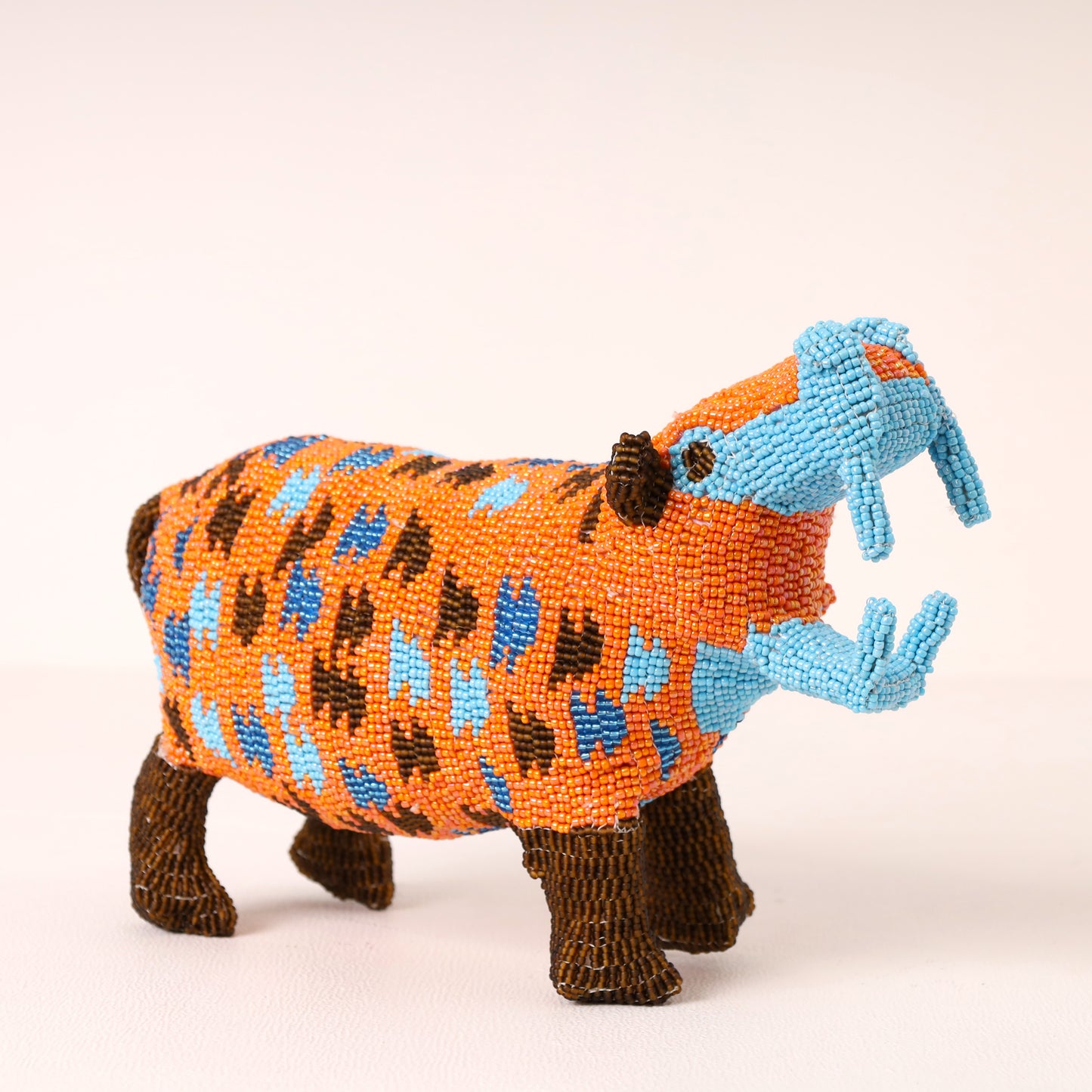 A handcrafted beaded hippopotamus figurine featuring an orange body with blue and brown spots, light blue tusks, and dark brown legs, set against a plain light background.