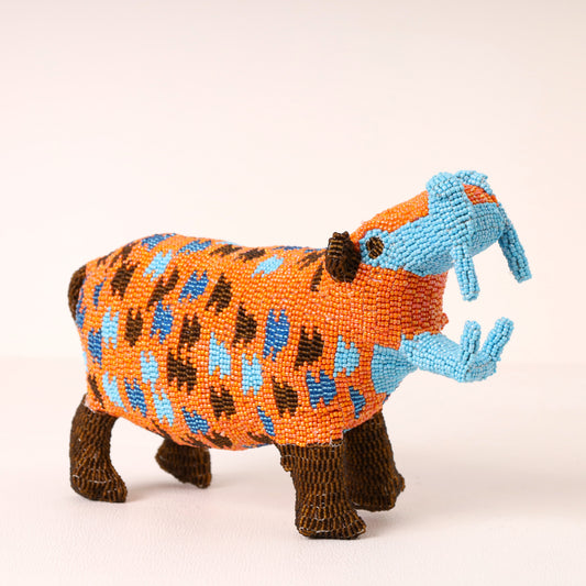 A handcrafted beaded hippopotamus figurine featuring an orange body with blue and brown spots, light blue tusks, and dark brown legs, set against a plain light background.