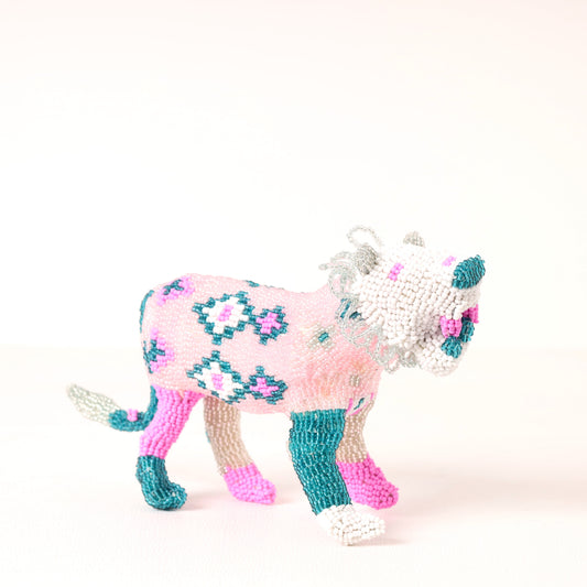 A handmade beaded lion figurine, decorated with a pink and white diamond pattern, teal accents, and pink and white hooves, set against a plain off-white background.