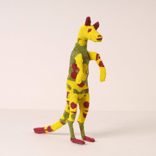 A handmade beaded meerkat figurine standing on two legs, featuring a yellow and green spotted pattern with red accents on its ears, eyes, hooves, and tail, set against a plain light beige background.