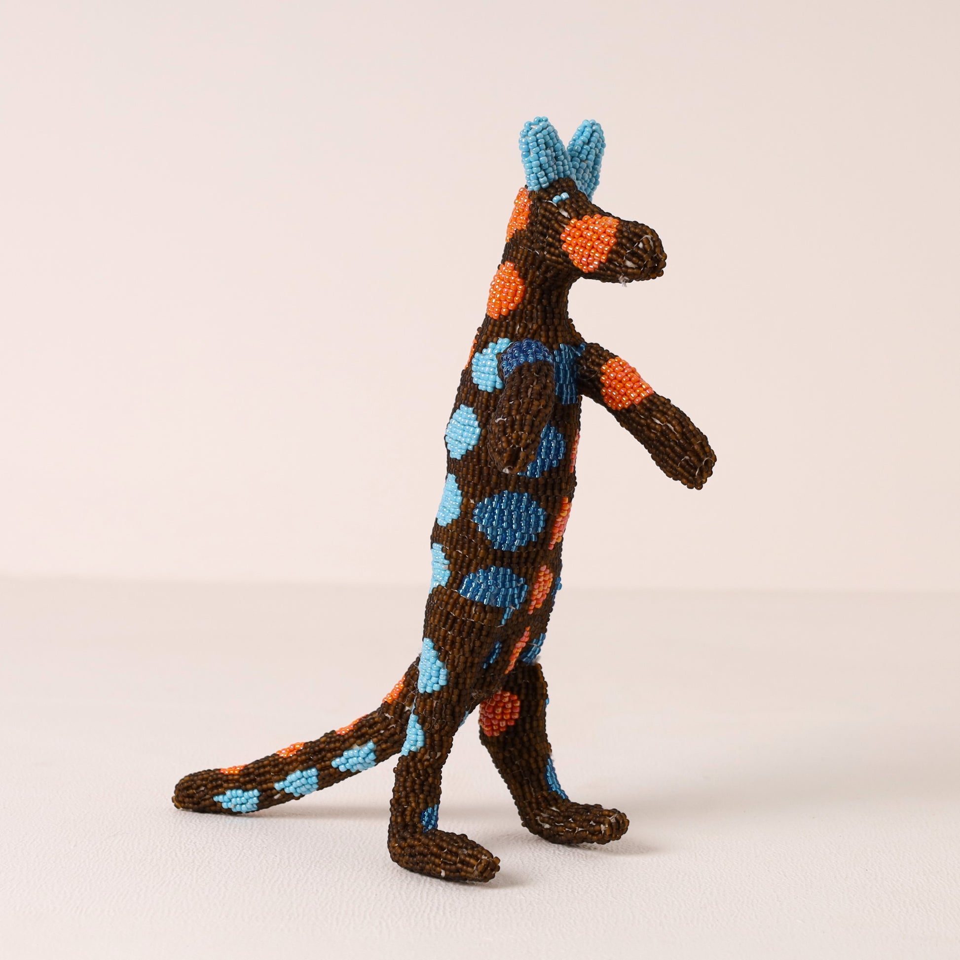 A handmade beaded meerkat figurine with bright blue and orange spots, standing on a light beige background.