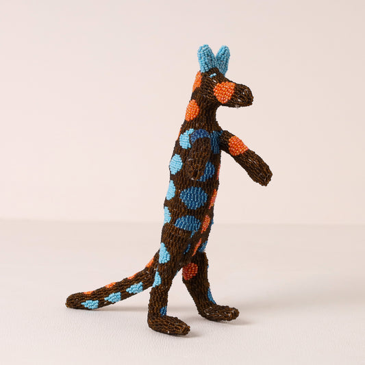 A handmade beaded meerkat figurine with bright blue and orange spots, standing on a light beige background.