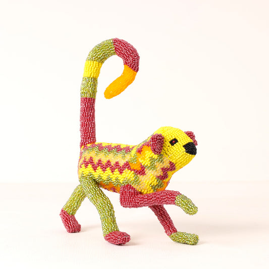 A handmade beaded monkey figurine, featuring a yellow body with red zigzag stripes, green legs and tail, and a yellow and red curved tail, set against a plain white background.