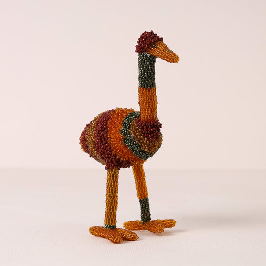 A handmade bead sculpture of an ostrich, crafted from multi-colored seed beads in rust orange, dark green, and deep red, standing on a light beige background.