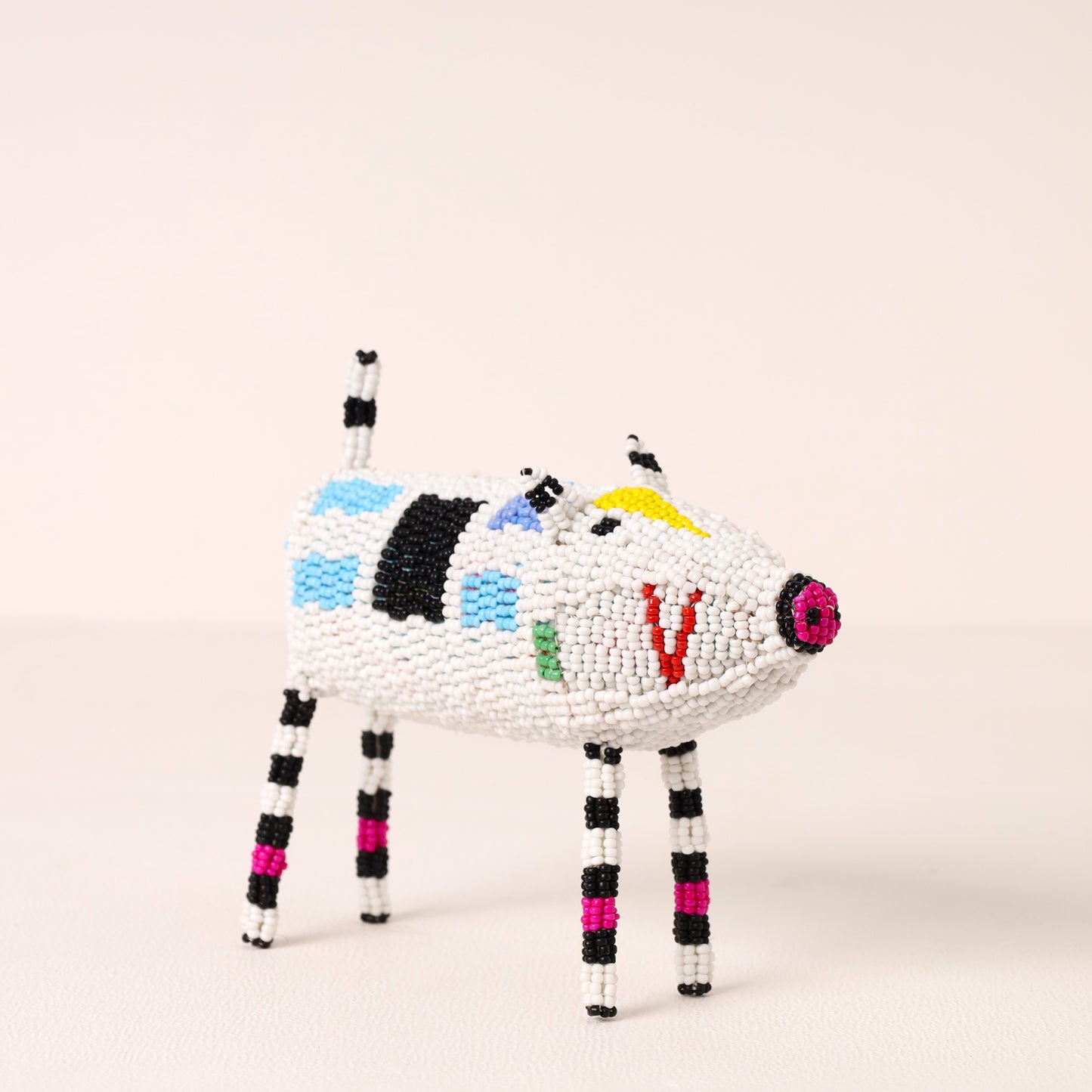 A handcrafted beaded animal figurine shaped like a pig, featuring a white body with black, blue, green, yellow, and red bead accents, and striped black and white legs, set against a plain light beige background.