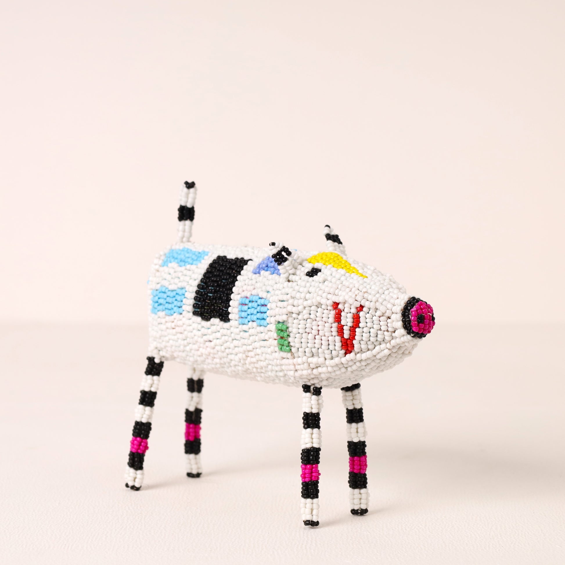 A handcrafted beaded animal figurine shaped like a pig, featuring a white body with black, blue, green, yellow, and red bead accents, and striped black and white legs, set against a plain light beige background.