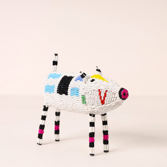 A handcrafted beaded animal figurine shaped like a pig, featuring a white body with black, blue, green, yellow, and red bead accents, and striped black and white legs, set against a plain light beige background.