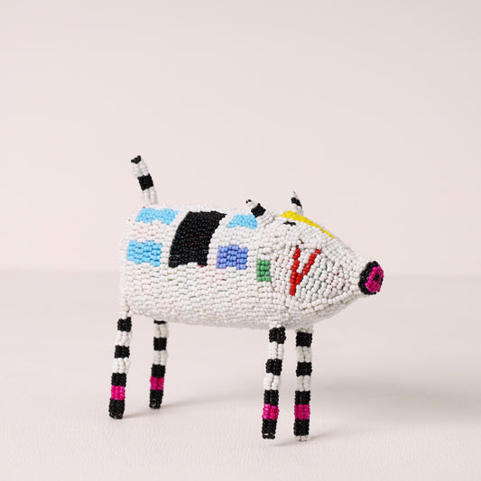 A handmade beaded pig figurine, crafted with white seed beads and decorated with colorful geometric patterns in black, blue, green, red, yellow, and pink, set against a plain light beige background.