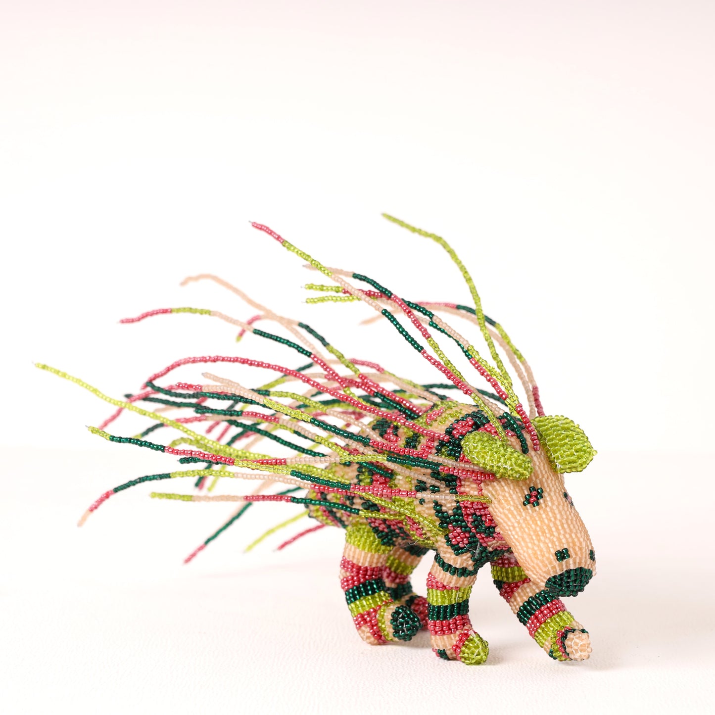 A handmade beaded porcupine figurine, featuring a beige body with green, pink, and black beadwork and long, wispy green and pink beaded tail-like extensions, photographed on a clean white background.