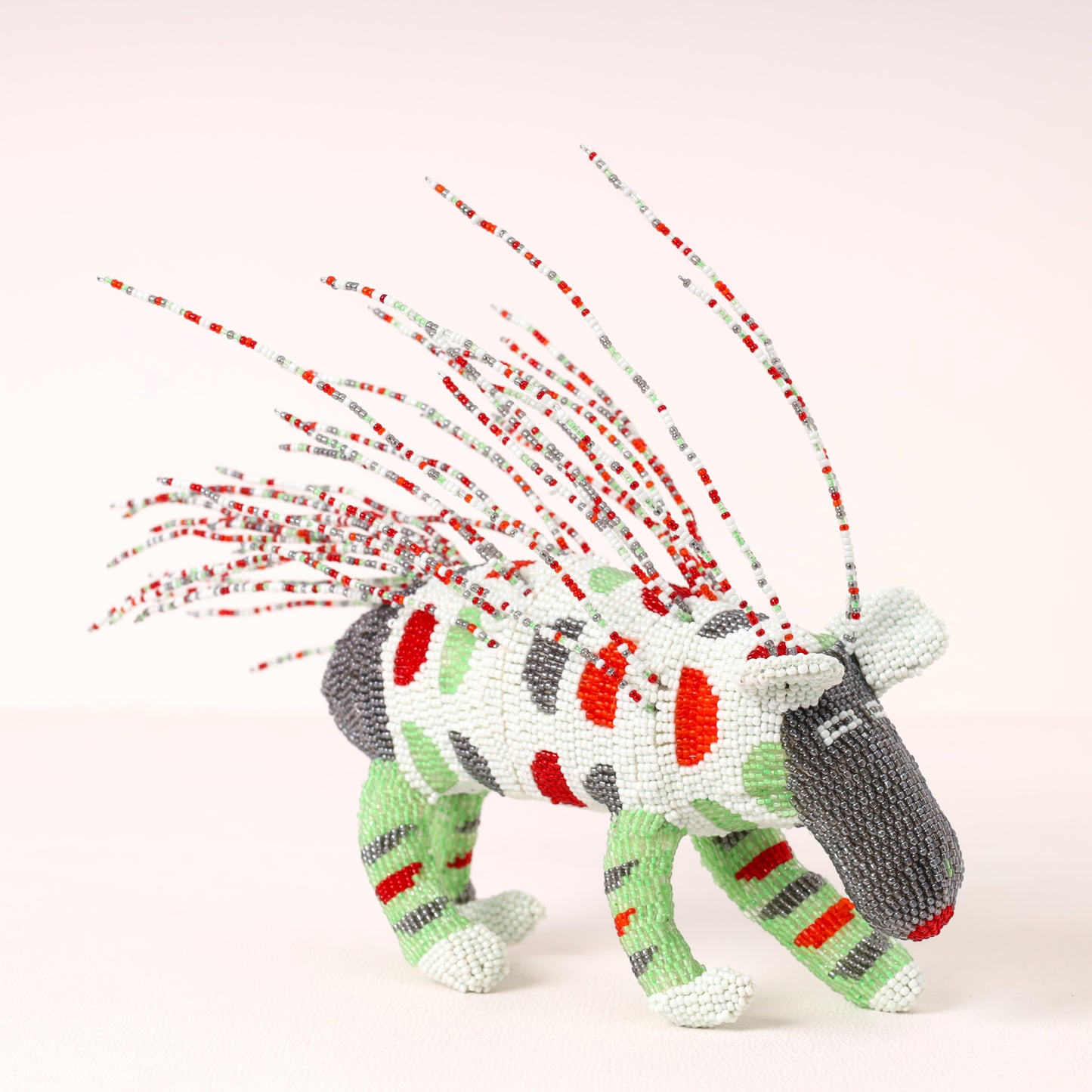 A handmade beaded porcupine sculpture with a white body, red and green stripes, gray head and back, and long red, white, and gray beaded mane spikes, photographed on a clean off-white background.
