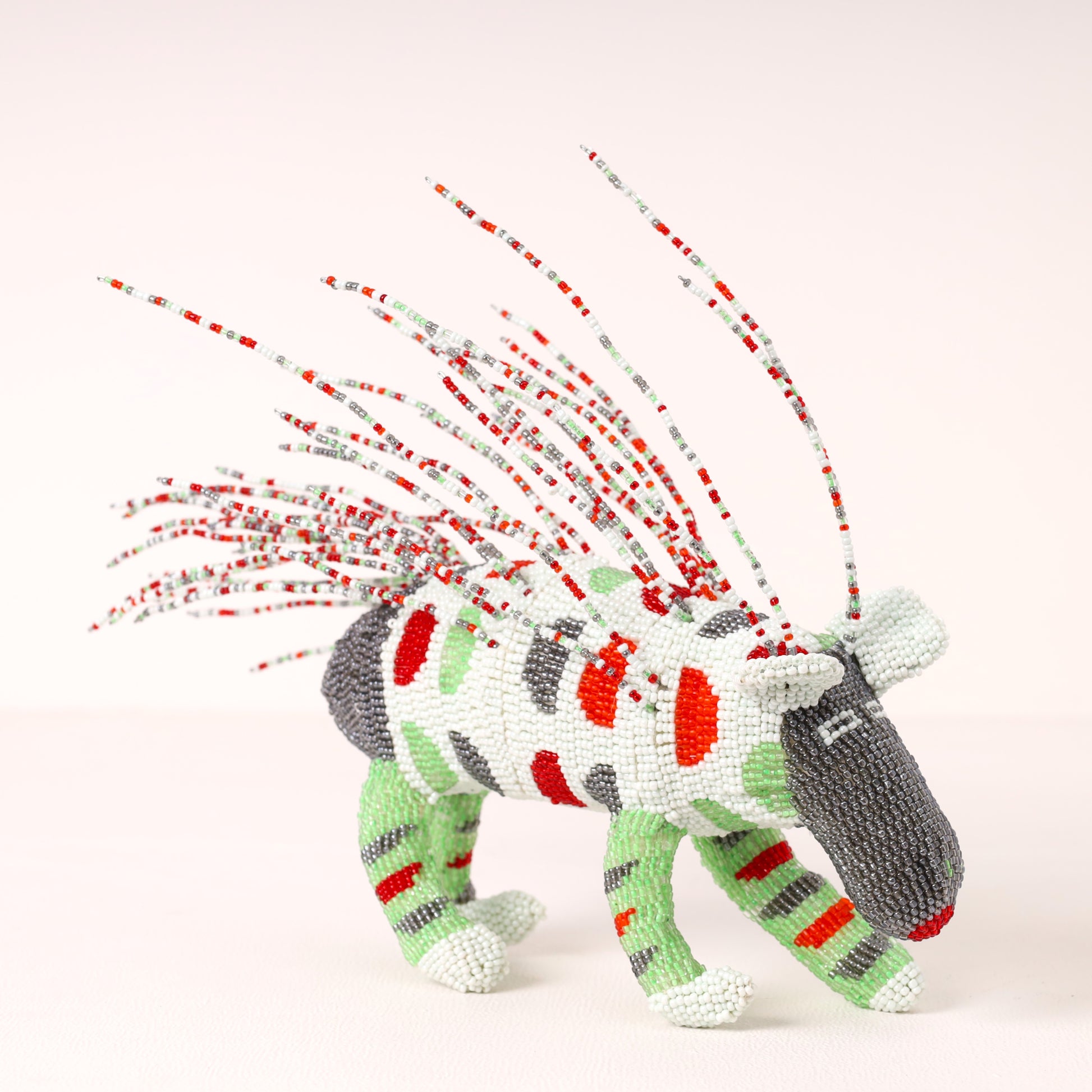 A handmade beaded porcupine sculpture with a white body, red and green stripes, gray head and back, and long red, white, and gray beaded mane spikes, photographed on a clean off-white background.