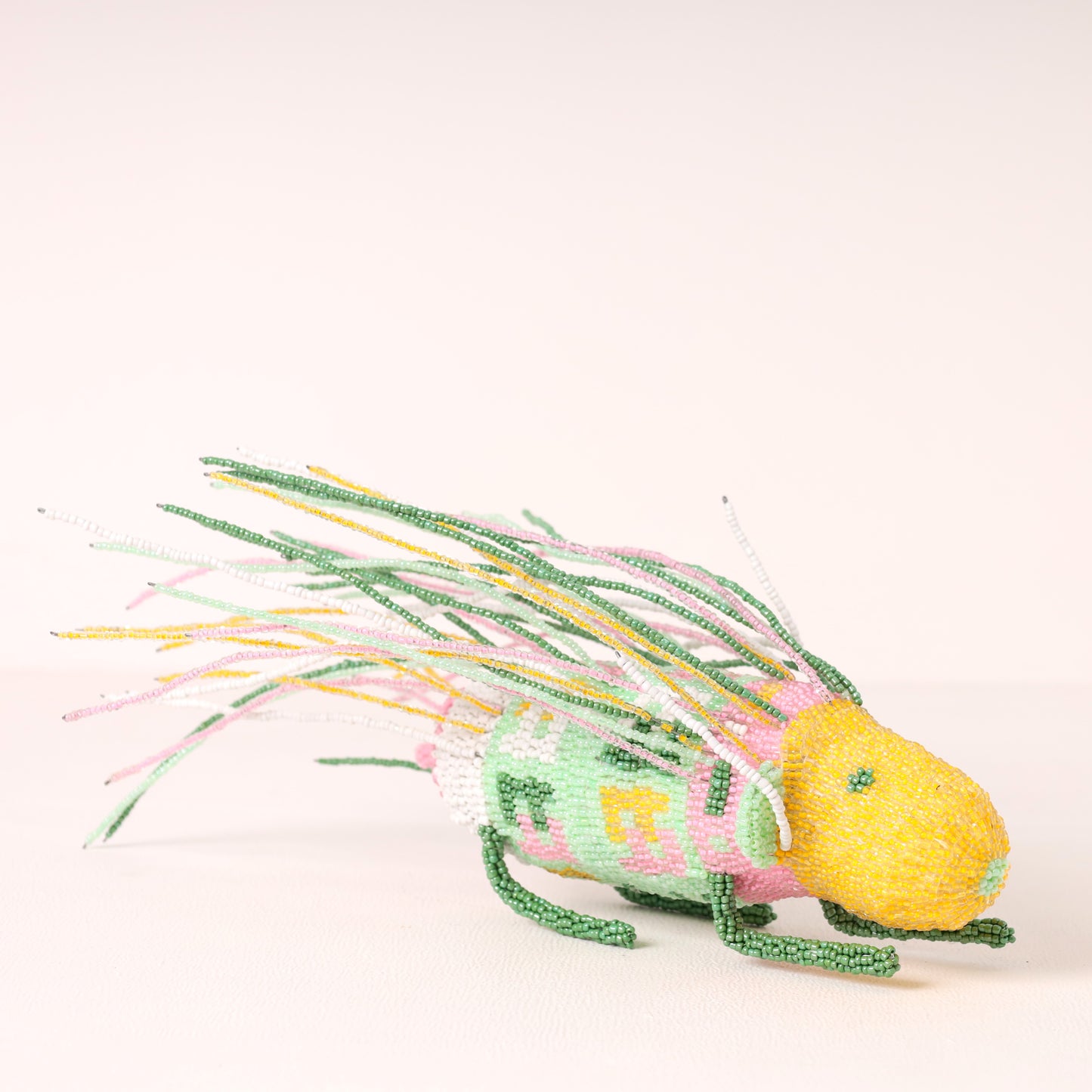 A handmade beaded porcupine ornament or decorative figure with a yellow head, green body, and multicolored beaded fins and tail on a plain light background.