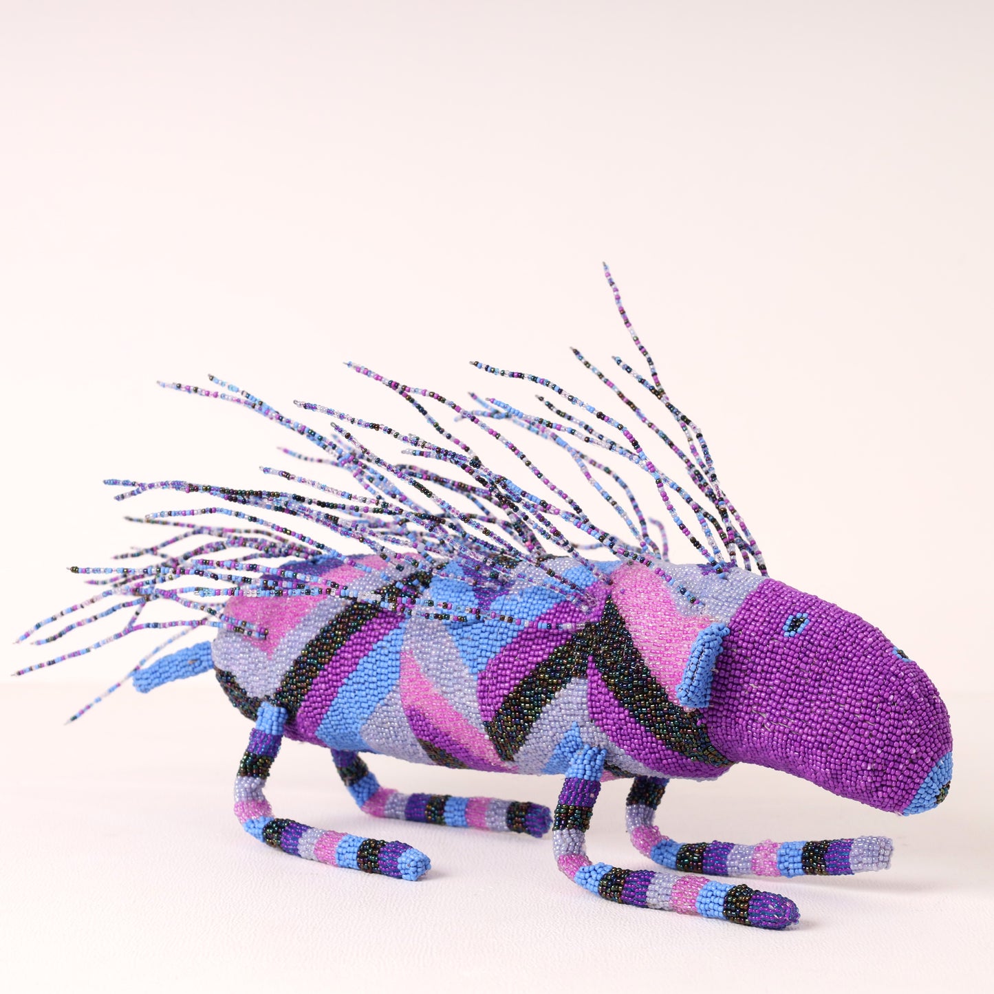 A handmade beaded porcupine sculpture crafted with purple, pink, blue, and black beads, featuring long spiky antennae, set against a plain light beige background.