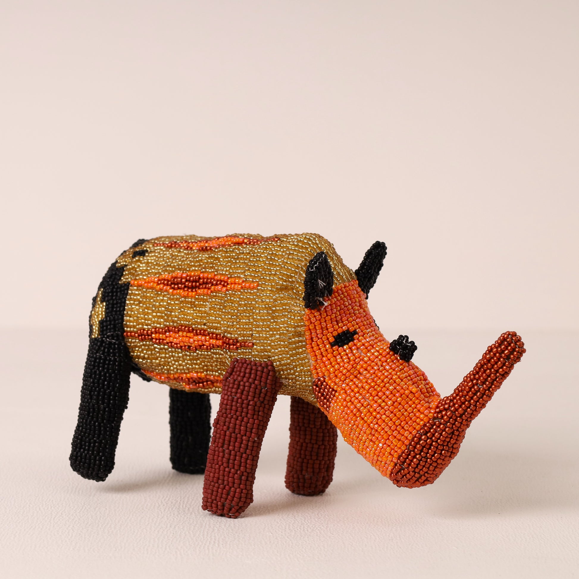 A handmade beaded rhinoceros figurine with a patterned body in gold, orange, and black, and a bright orange snout and legs, standing on a light beige surface against a neutral background.