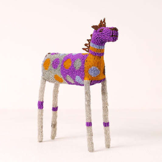 A handmade beaded zebra figurine with a purple head and neck, a patterned body of purple, orange, and gray, and textured gray legs with purple accents, set against a plain light beige background.
